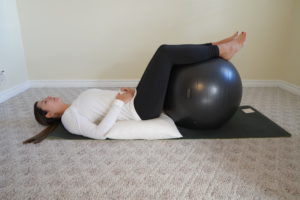 Laura, laying on her back on a black yoga mat. There is a white pillow under her pelvis and back. Her legs are up on a grey exercise ball. Her hands are resting on her pelvis. She is looking at the ceiling. 
