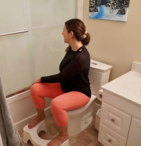 Laura in pink leggings and a black shirt. She is sitting on a white toilet with her feet on a white stool.