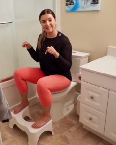 Laura in pink leggings and a black shirt. She is sitting on a white toilet with her feet on a white stool. She is point to the stool and smiling. 