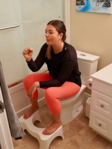 Laura in pink leggings and a black shirt with her feet on a stool. She is blowing into a fist and leaning forward.