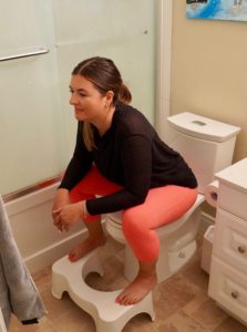 Laura in pink leggings and a black shirt. She is sitting on a while toilet with her feet on a white stool and is leaning forward. 